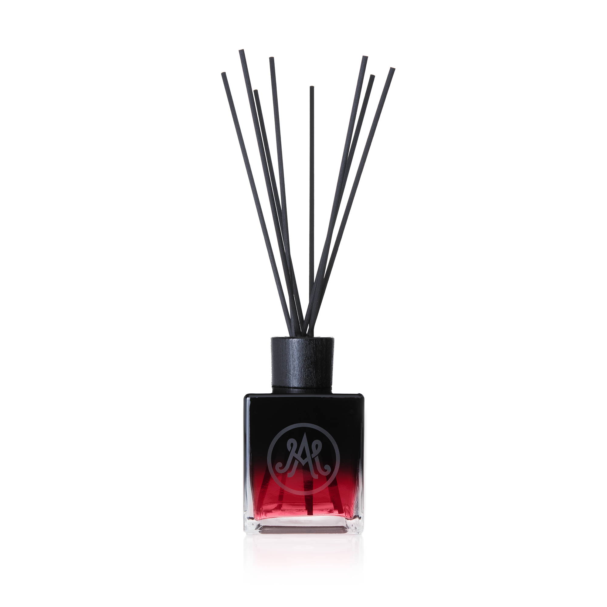 A dark red diffuser with black reeds is displayed, featuring a sleek black cap and an elegant monogram logo on the front of the bottle. Sweet fig blends with the aroma of warm grass and ripe plums.